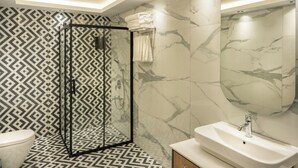 A modern bathroom with a geometric patterned floor, a glass shower enclosure, a marble-tiled wall, and a white sink. - Yeşilton Otel (Arhavi)
