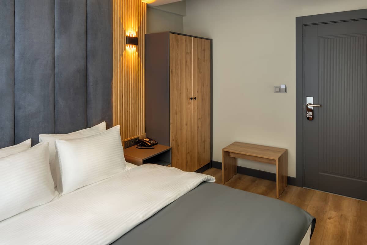 A modern hotel room with a large bed, a wardrobe, a bedside table with a phone, and a small wooden bench.
