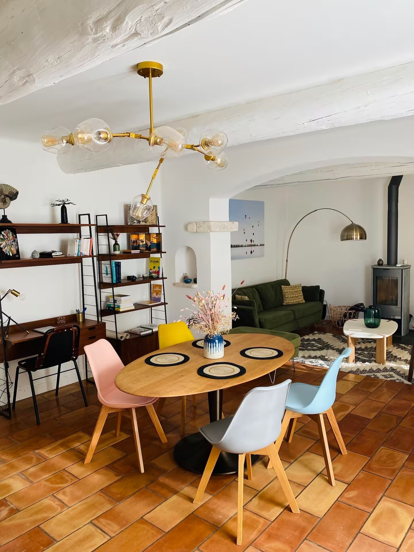 A modern living room with a wooden dining table, colorful chairs, a bookshelf, a green sofa, and a fireplace.