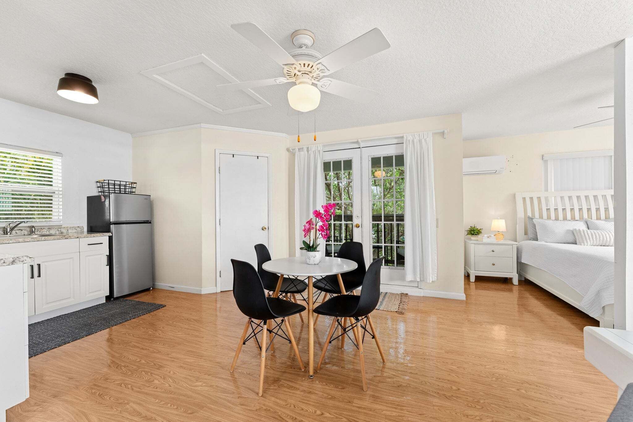 A modern kitchen with a refrigerator, a dining table with chairs, a ceiling fan, and a bed with a white headboard.