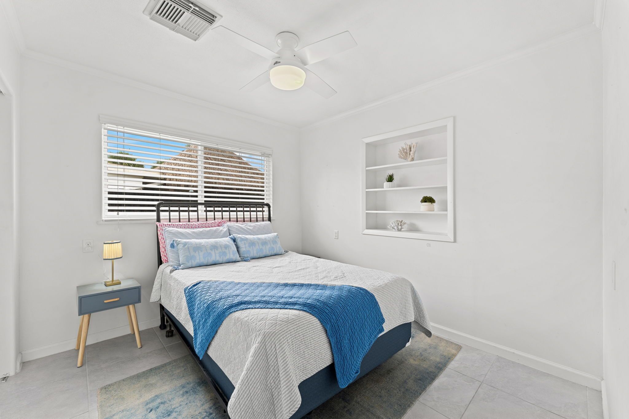 Quarto com cama, mesa de cabeceira, luminária, janela com persianas, ventilador de teto e prateleira na parede com objetos decorativos.