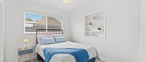 Quarto com cama, mesa de cabeceira, luminária, janela com persianas, ventilador de teto e prateleira na parede com objetos decorativos.