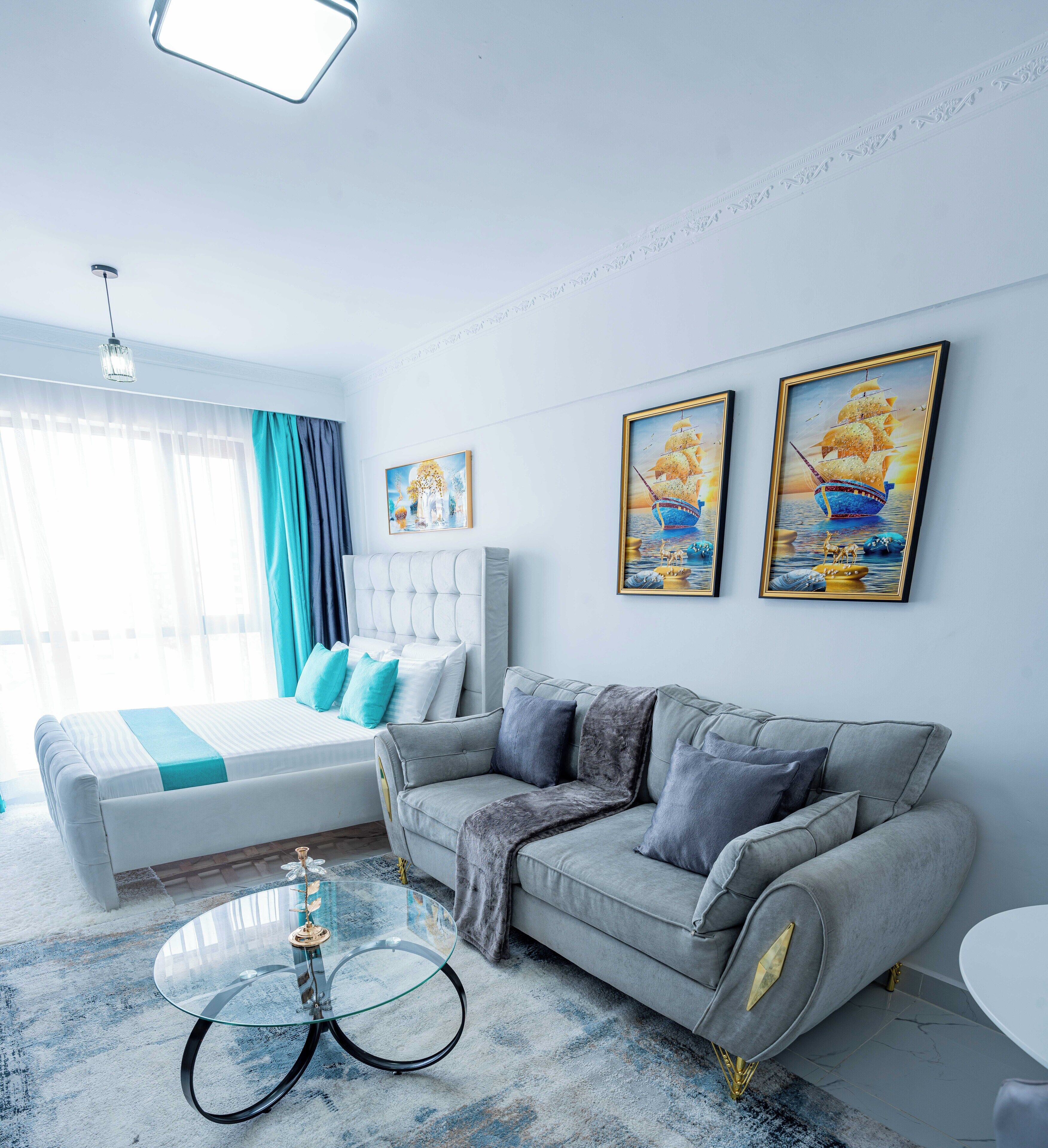 A modern bedroom with a bed, a grey sofa, a glass coffee table, and two framed paintings on the wall.