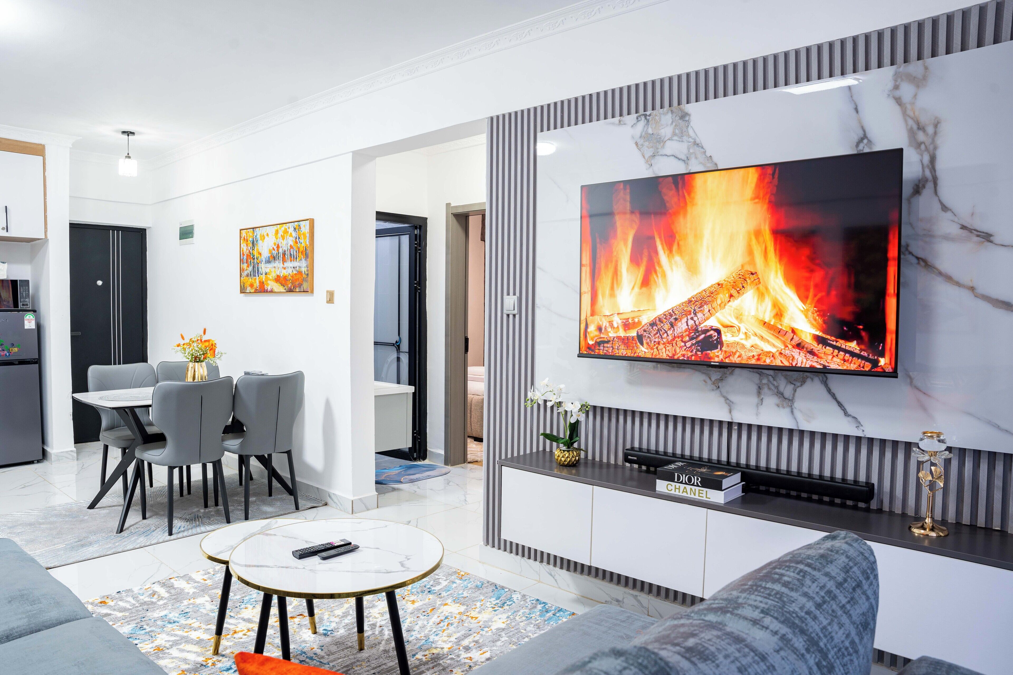 A modern living room with a flat-screen TV, grey sofas, a dining area with a table and chairs, and a kitchen in the background.