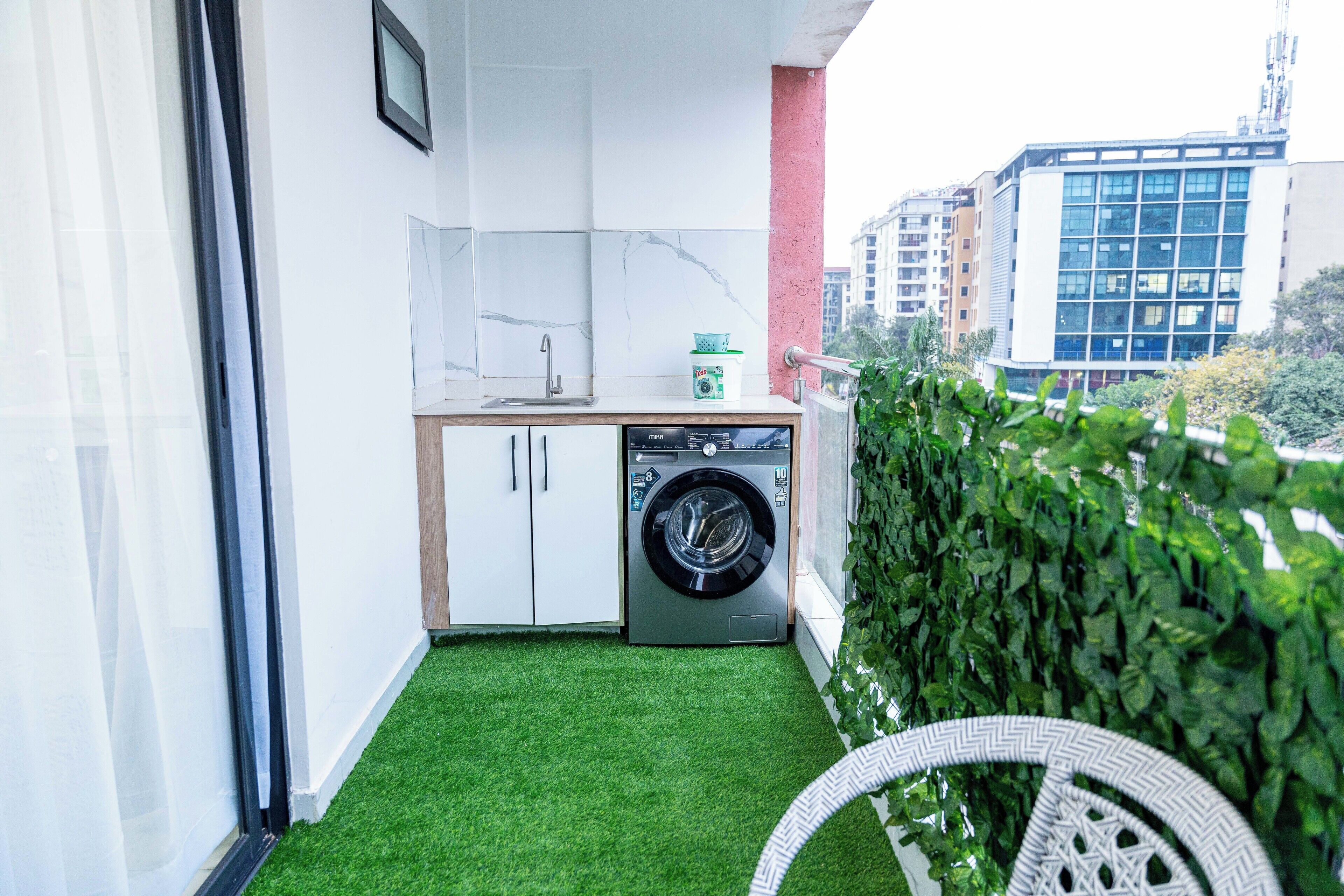Un petit balcon agrémenté d’un gazon synthétique, d’un lave-linge et d’un coin cuisine avec des meubles blancs et un évier.