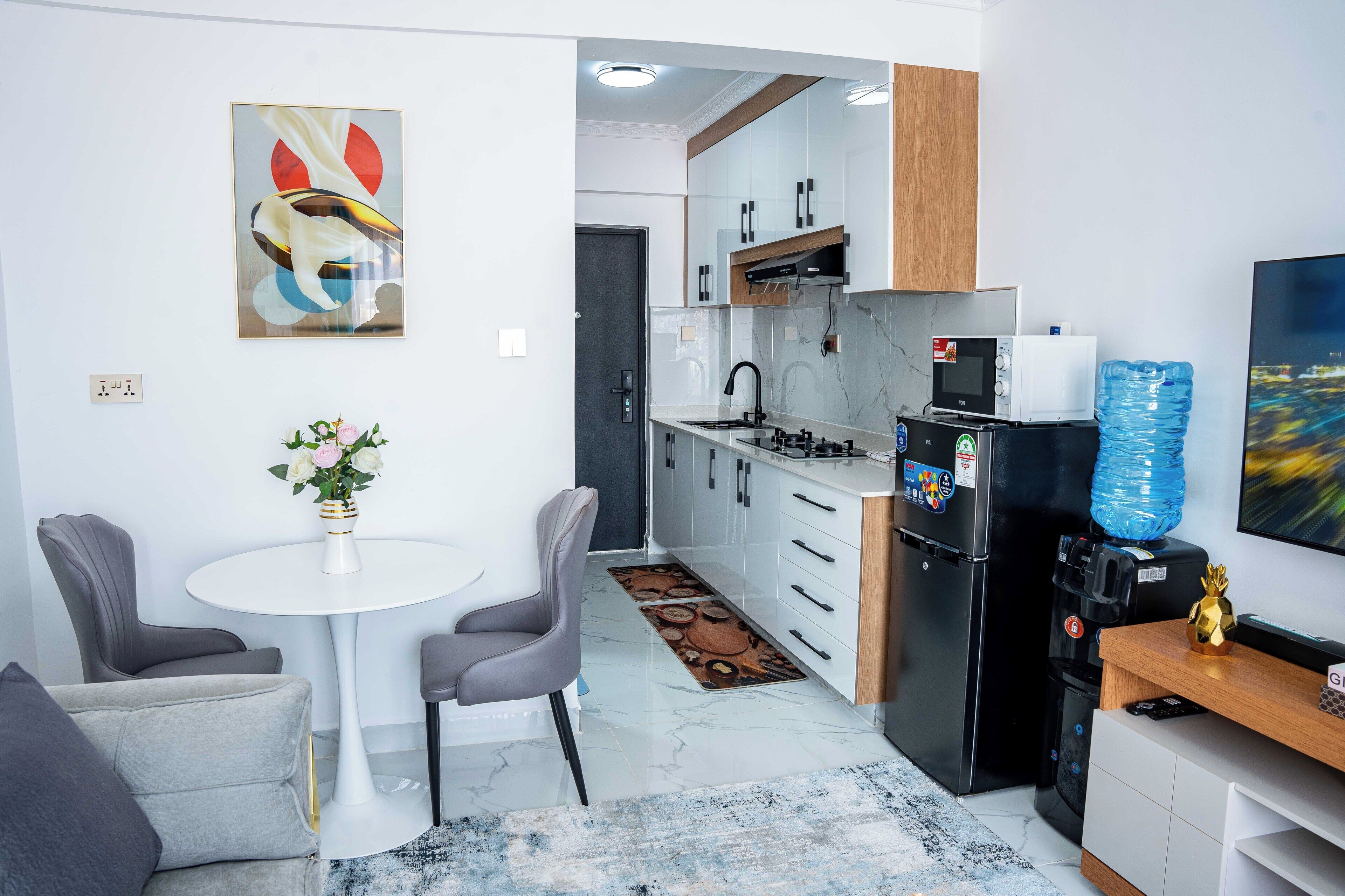 A modern kitchen with white cabinets, a black refrigerator, and a water dispenser. There is a dining area with a white table and two chairs.
