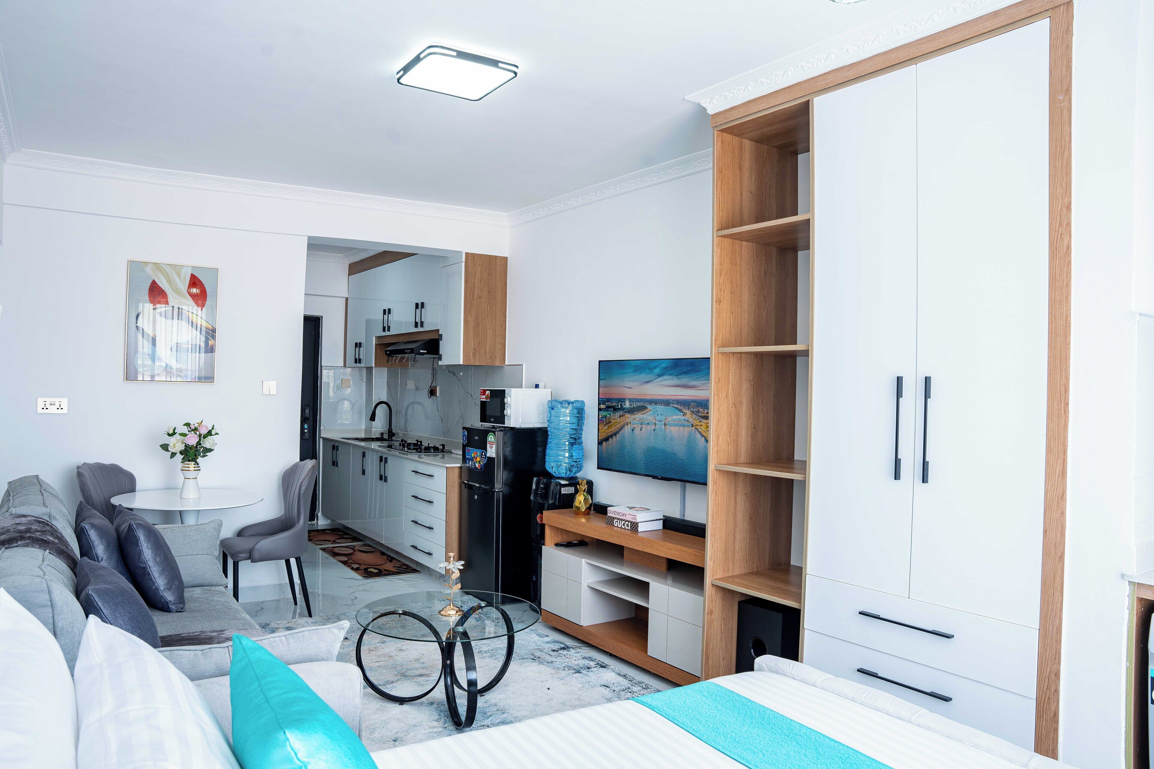 A modern living room with a sofa, coffee table, and TV. There is a kitchen area with cabinets and a refrigerator.