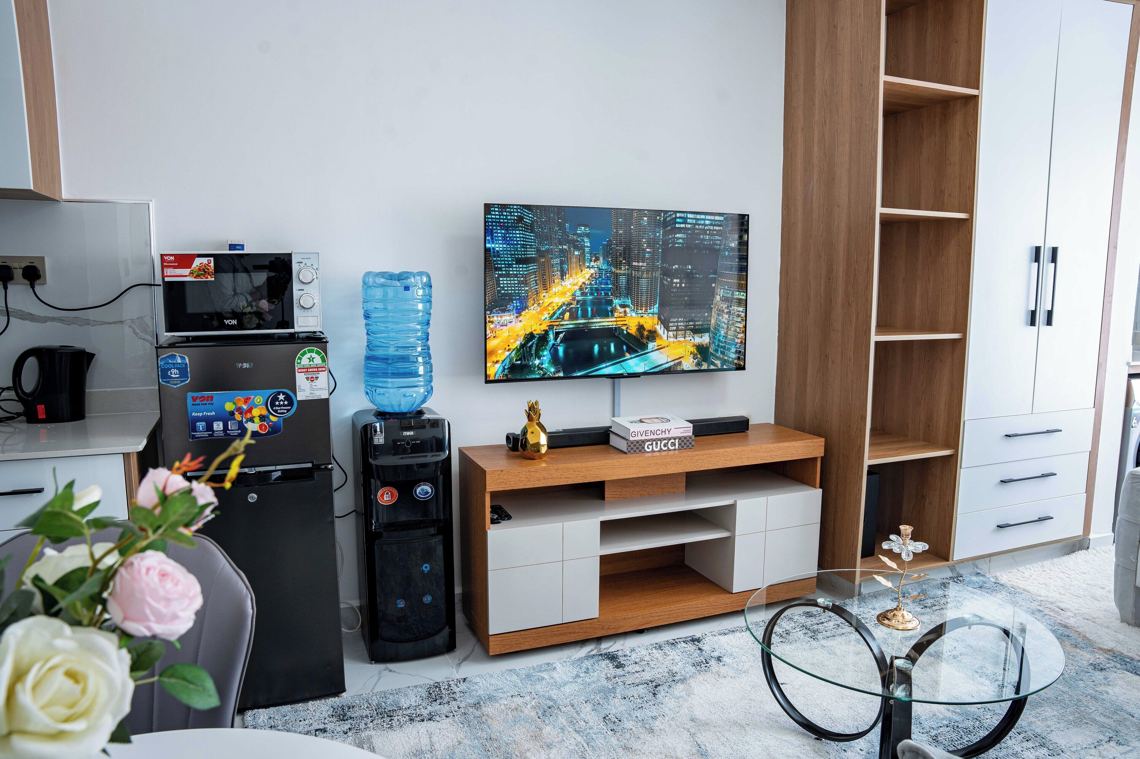A modern hotel room with a flat-screen TV, a mini-fridge, a water dispenser, a coffee maker, a glass table, and a sofa.