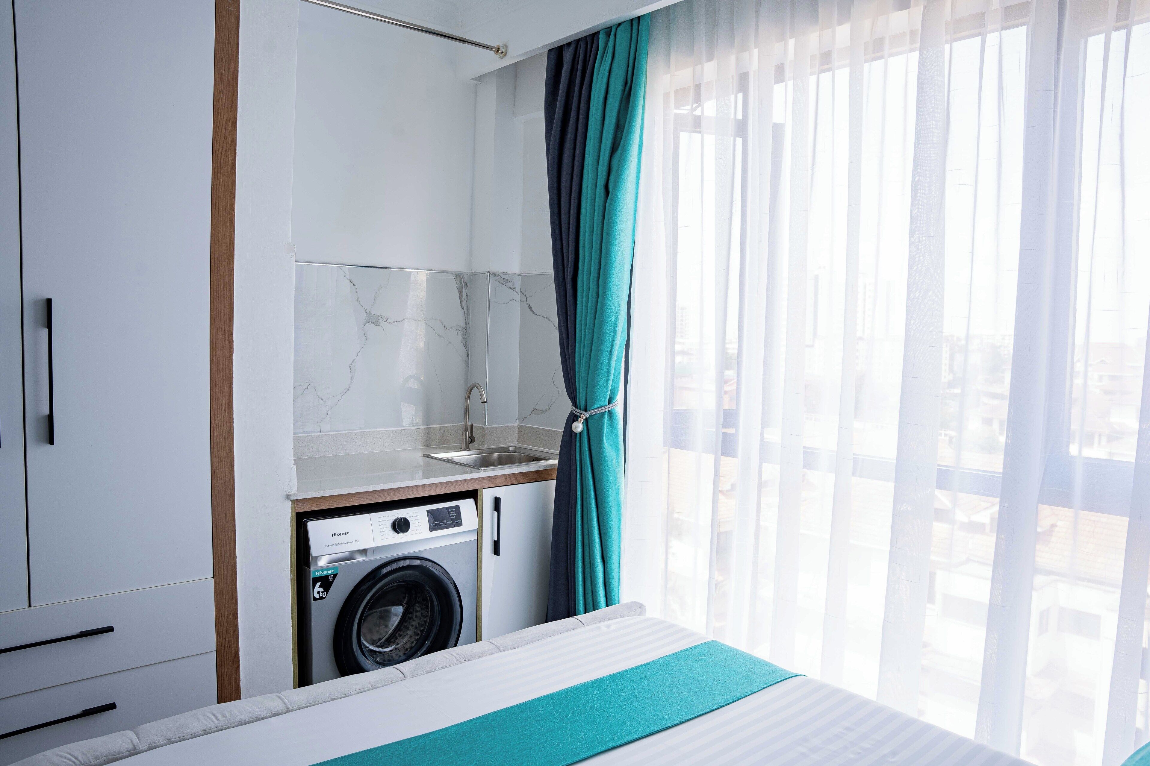 Een kleine hotelkamer met een wasmachine, een bed en uitzicht op de stad door dunne gordijnen.