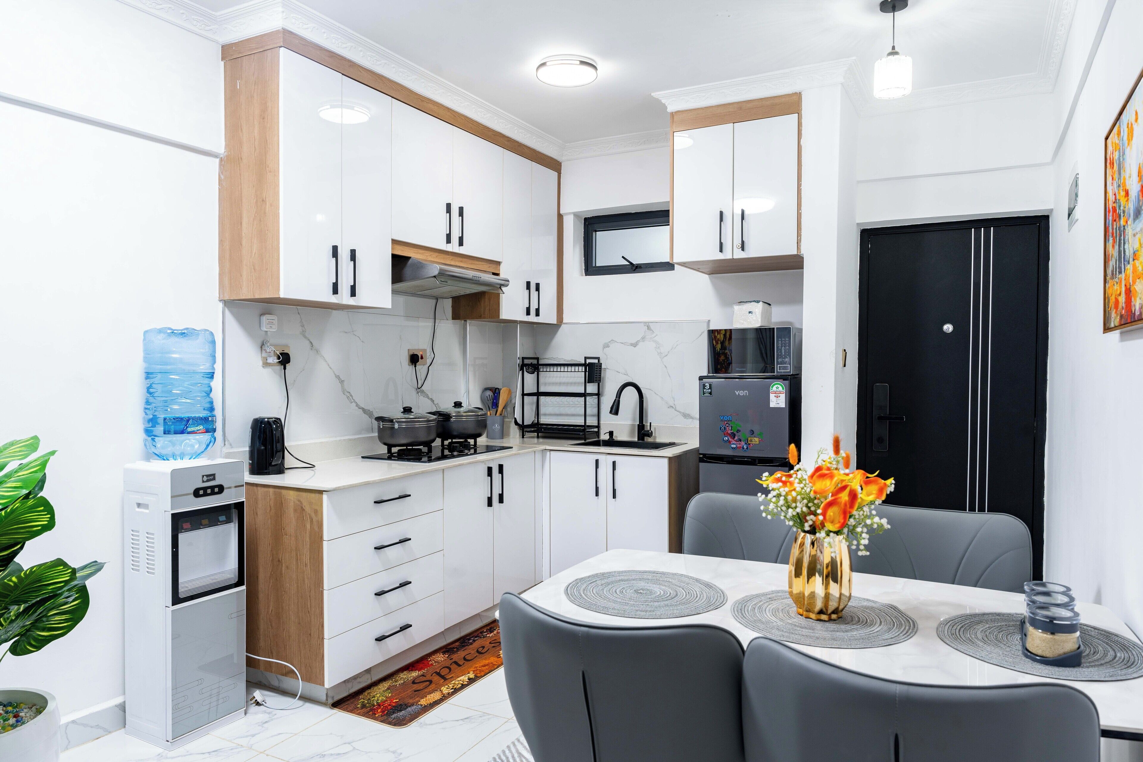 A modern kitchen with white cabinets, a water dispenser, a microwave, and a refrigerator. There is a dining table with chairs and a vase with flowers.