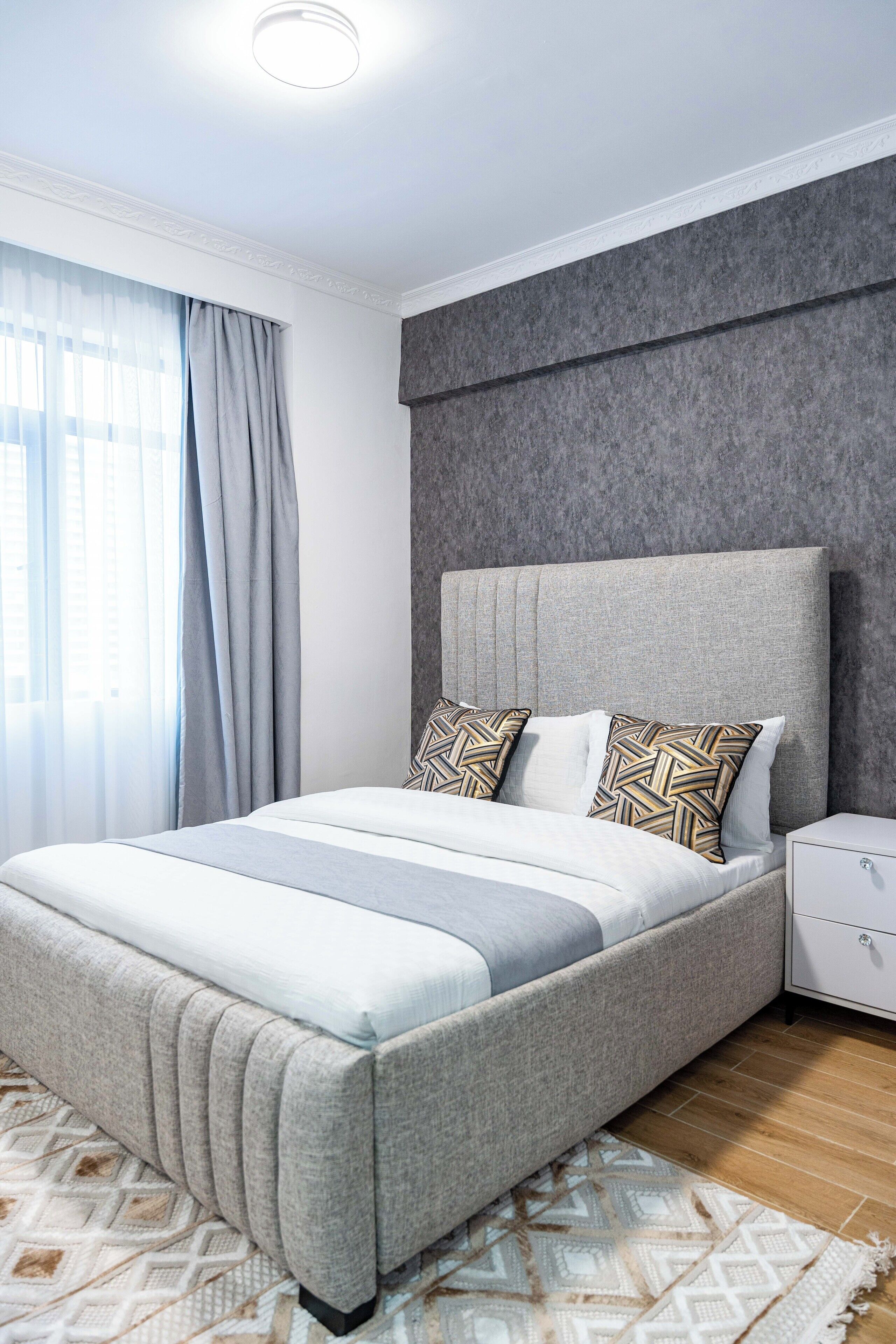 A modern bedroom with a large bed, grey upholstered headboard, and patterned pillows.