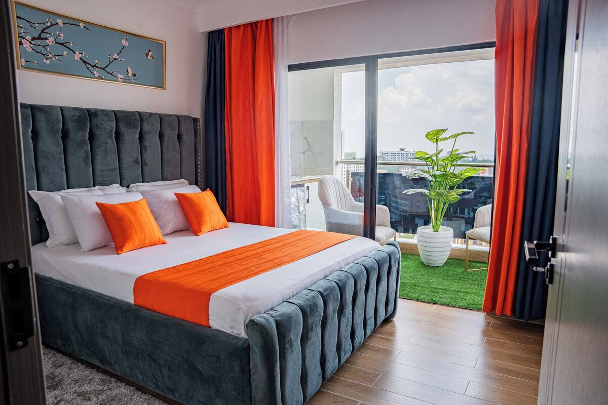 A modern bedroom with a large bed, a view of the outdoors, and a balcony with a potted plant.