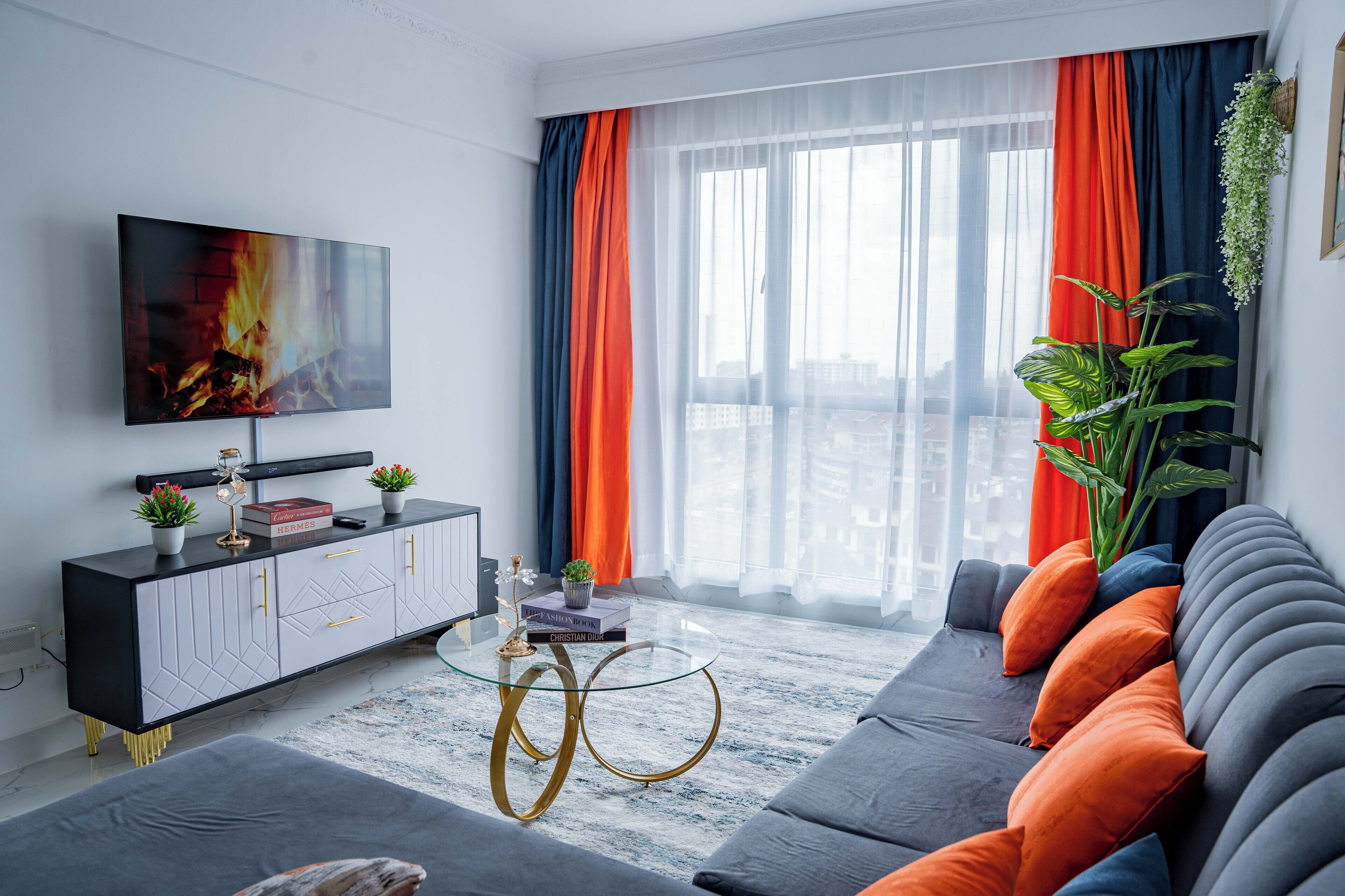 Un salon moderne doté d’un grand téléviseur à écran plat, d’un meuble noir et blanc, d’une table basse en verre et d’un canapé gris agrémenté de coussins orange.