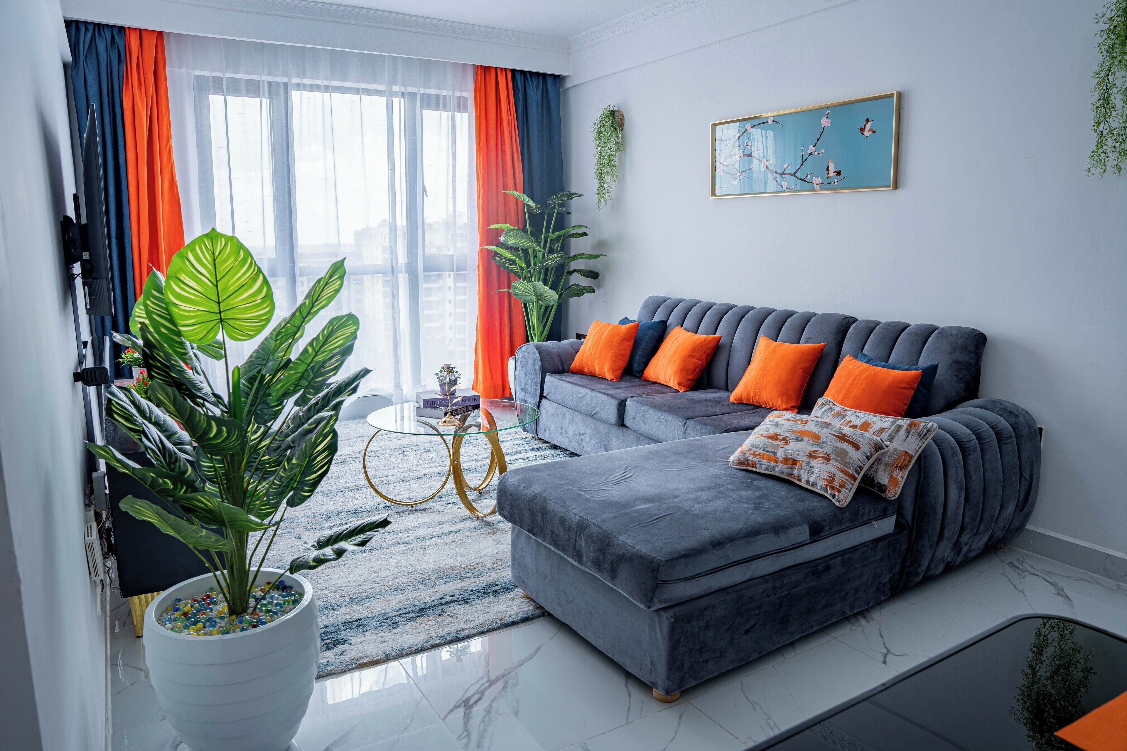 Un salón moderno con un sofá gris, cojines anaranjados, una planta grande en maceta y un cuadro en la pared.