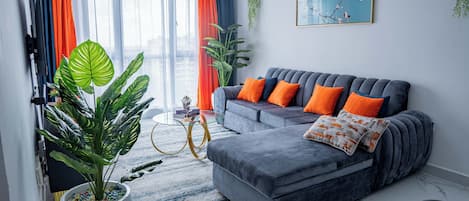 Un salón moderno con un sofá gris, cojines anaranjados, una planta grande en maceta y un cuadro en la pared.