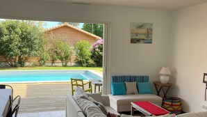 A modern living room with a sofa, a dining table, and chairs. There is a view of a swimming pool and a wooden building in the background. - 4-bedroom villa, private heated pool, enclosed garden8 minutes from the beach (Linxe)