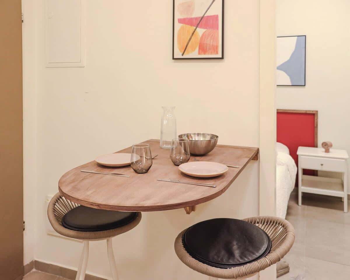 A dining area with a round wooden table, two wicker chairs with black cushions, and a white wall with abstract art.