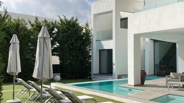 A modern outdoor pool area with lounge chairs and umbrellas, surrounded by greenery and a contemporary building.