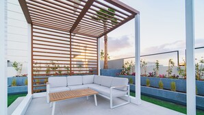 A modern outdoor seating area with a white sofa and wooden table, surrounded by blue planters and a wooden pergola. - Quiet Top-Floor Studio Near Metro + Baby Gear (Dubai)