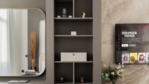 A modern hotel room with a large mirror, a built-in shelf with various items, a small table with a laptop, and a wall-mounted TV displaying a show. - Quiet Top-Floor Studio Near Metro + Baby Gear (Dubai)