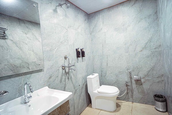 Un baño con lavamanos, inodoro y ducha de blanco, paredes de mármol y un cubo de basura.