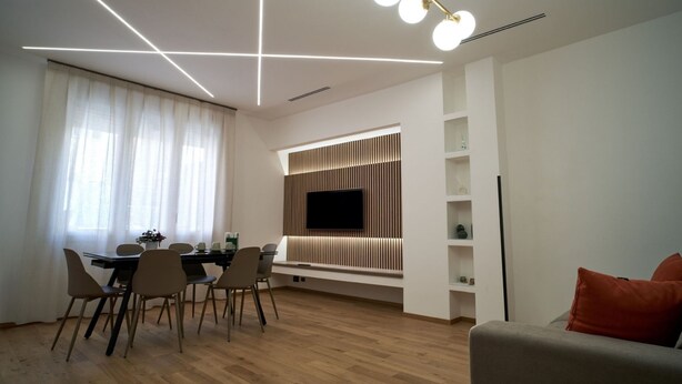 Een moderne woonkamer met een eethoek, houten vloeren, een flatscreen televisie bevestigd aan een houten wandpaneel, en een plafondlamp.