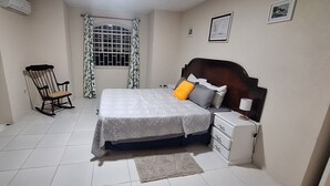 A bedroom with a bed, a nightstand, a chair, a fan, a window with curtains, and a wall-mounted air conditioner. - Cottage at Coverley (Christ Church)