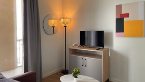 A modern living room with a sofa, a TV on a wooden stand, a floor lamp, a small table with a plant, and a painting on the wall. - Zenao Appart'hôtels Cognac (Cognac)