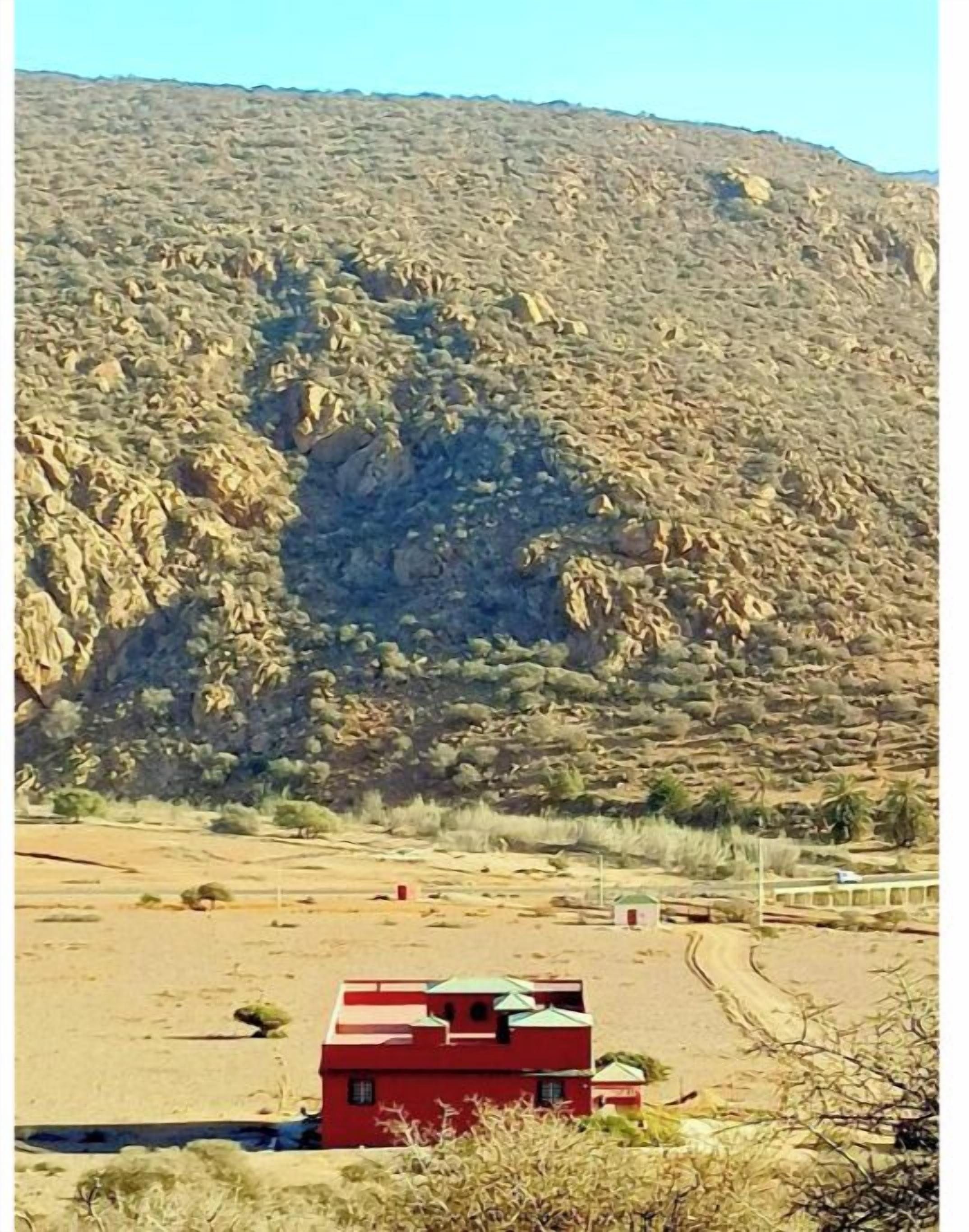 A red building with a green roof, surrounded by a dry landscape and a rocky hill in the background.
