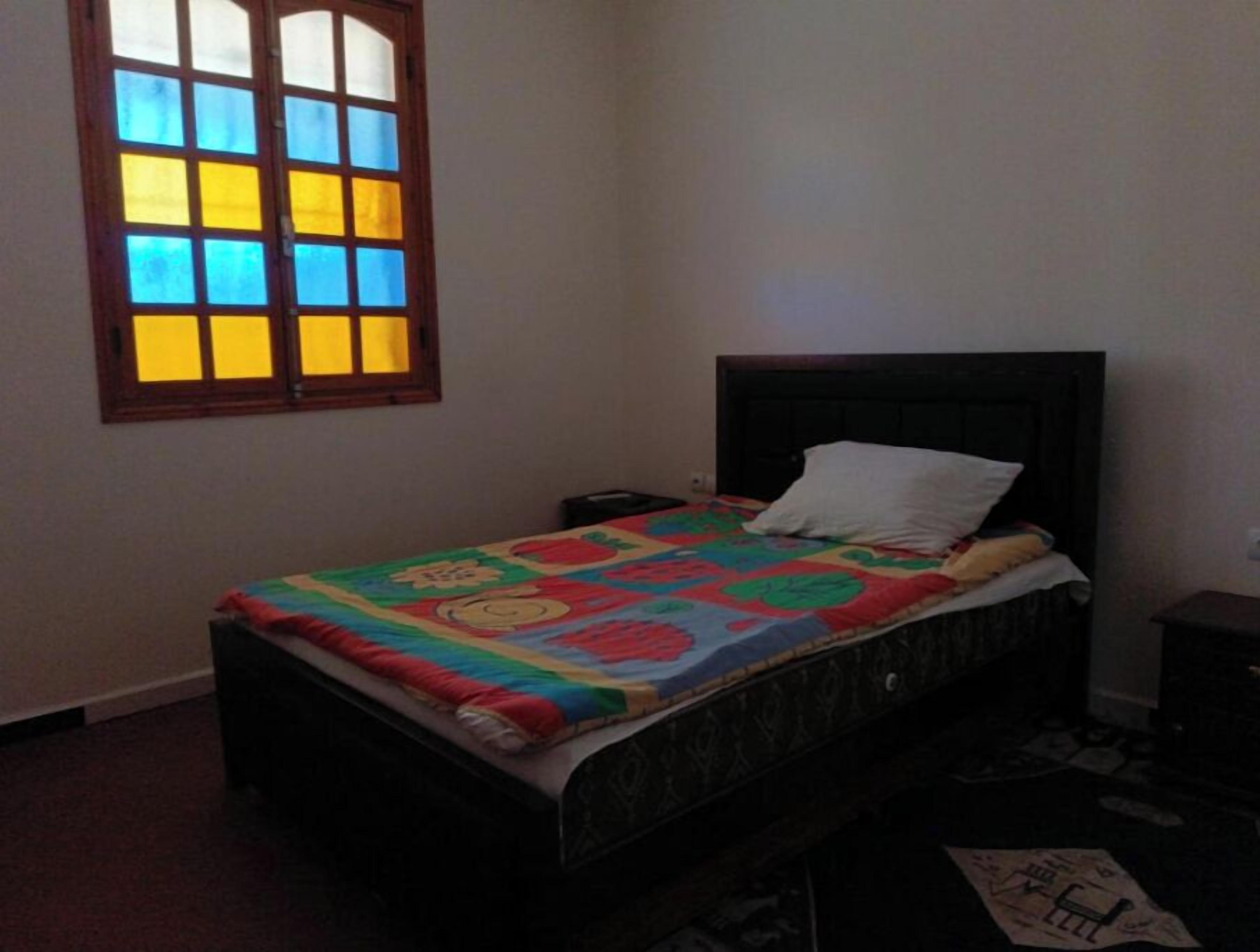 A bedroom with a bed, a nightstand, a window with stained glass, and a wall-mounted shelf.