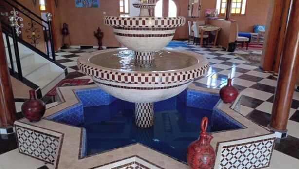 A multi-tiered fountain in a lobby with checkered floor tiles, a staircase, and a dining area. - Auberge Assif iboudrarn (Tioughza)