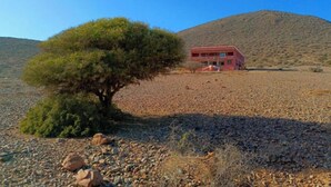 A red building with a tree in front, set in a rocky landscape with a hill in the background. - Auberge Assif iboudrarn (Tioughza)