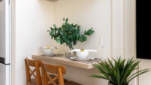 Eine Küche mit einem Holztisch, Stühlen, einem weißen Kühlschrank, einem Topf mit Pflanze und einem an der Wand montierten Flachbildfernseher.