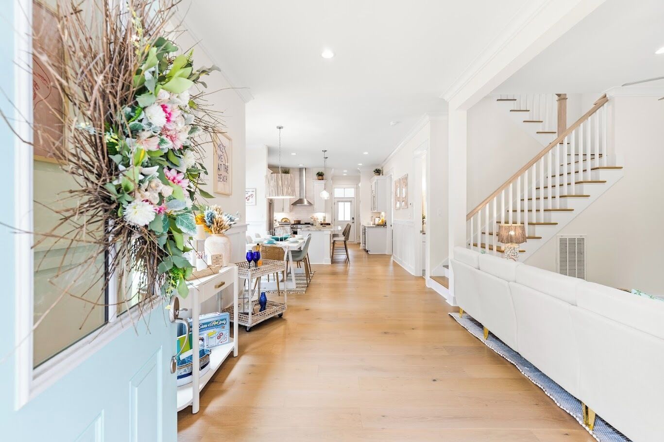 A spacious hallway with a wooden floor, a white staircase, and a dining area in the background.