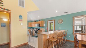 A kitchen with wooden cabinets, a central island with bar stools, a microwave, and a clock on the wall. - Beach Therapy: Private Heated Pool and Hot Tub (Corolla)