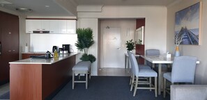 Une cuisine moderne avec des meubles blancs, une machine à café et un évier. On y trouve un coin repas avec une table et des chaises, ainsi qu’une grande plante en pot.