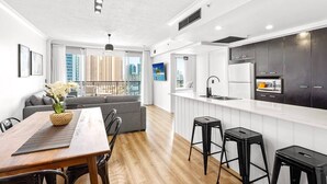 A modern kitchen with white cabinets, a black countertop, and a built-in oven. There is a dining area with a wooden table and chairs, and a gray sofa. - Chevron With 3beds and own Carpark Within Tower3 (Gold Coast)