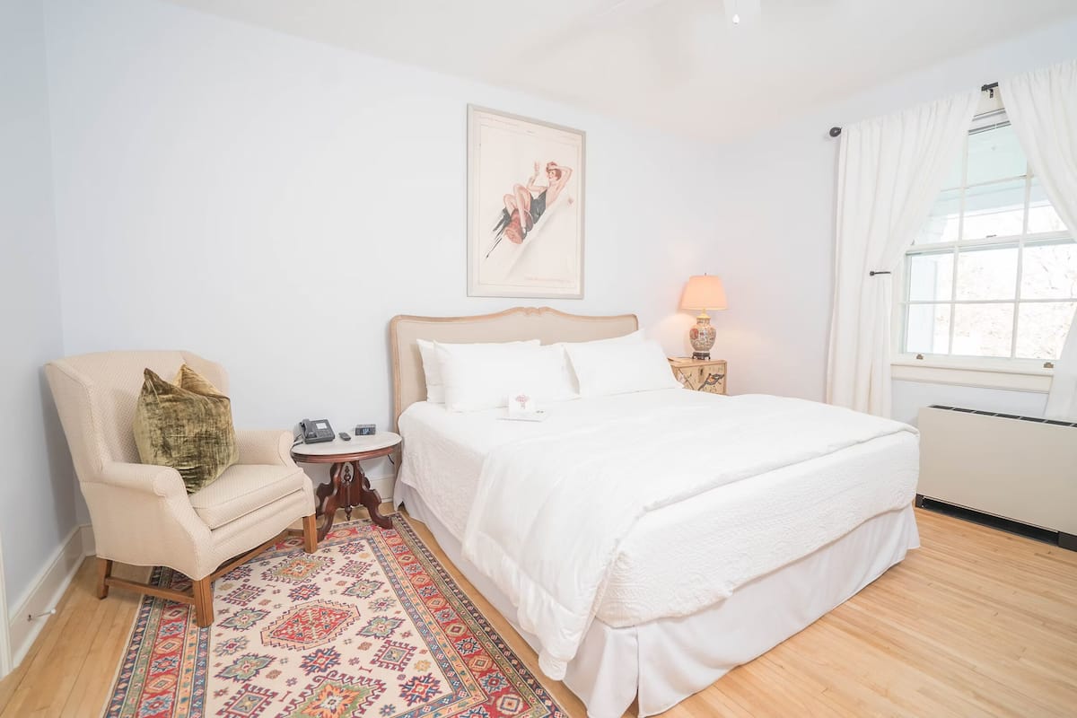 A bedroom with a large bed, a chair, a small table, a lamp, and a window with curtains.
