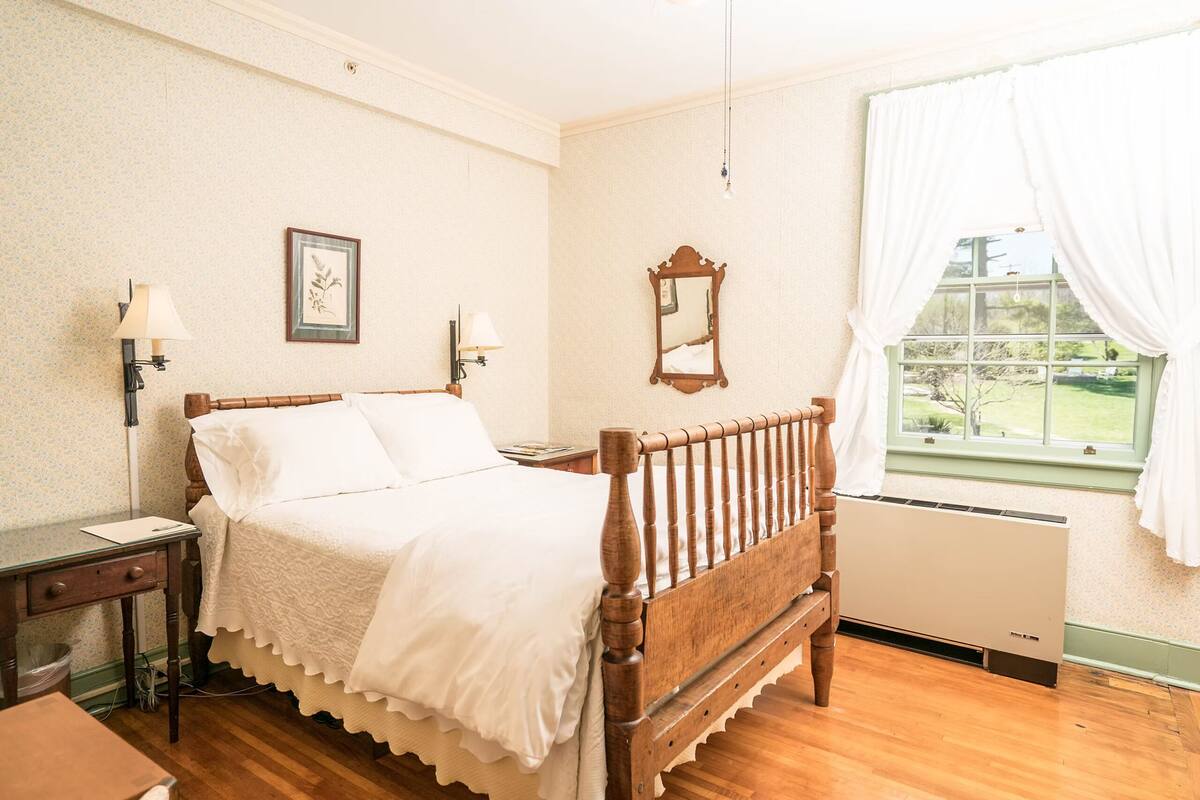 A bedroom with a wooden bed, a nightstand, a desk, a radiator, and a window with curtains.