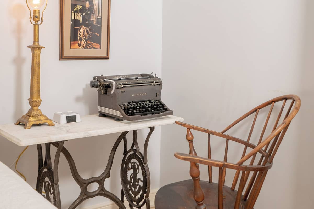 A corner room with a bed, a desk with a typewriter, a wooden chair, a lamp, and a framed picture on the wall.