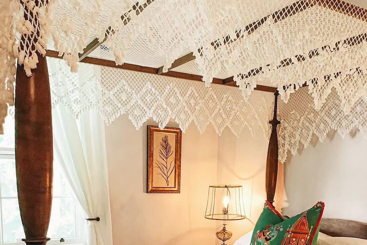 A four-poster bed with a lace canopy, a framed botanical print on the wall, a bedside table with a lamp, and a green pillow with a Christmas theme.