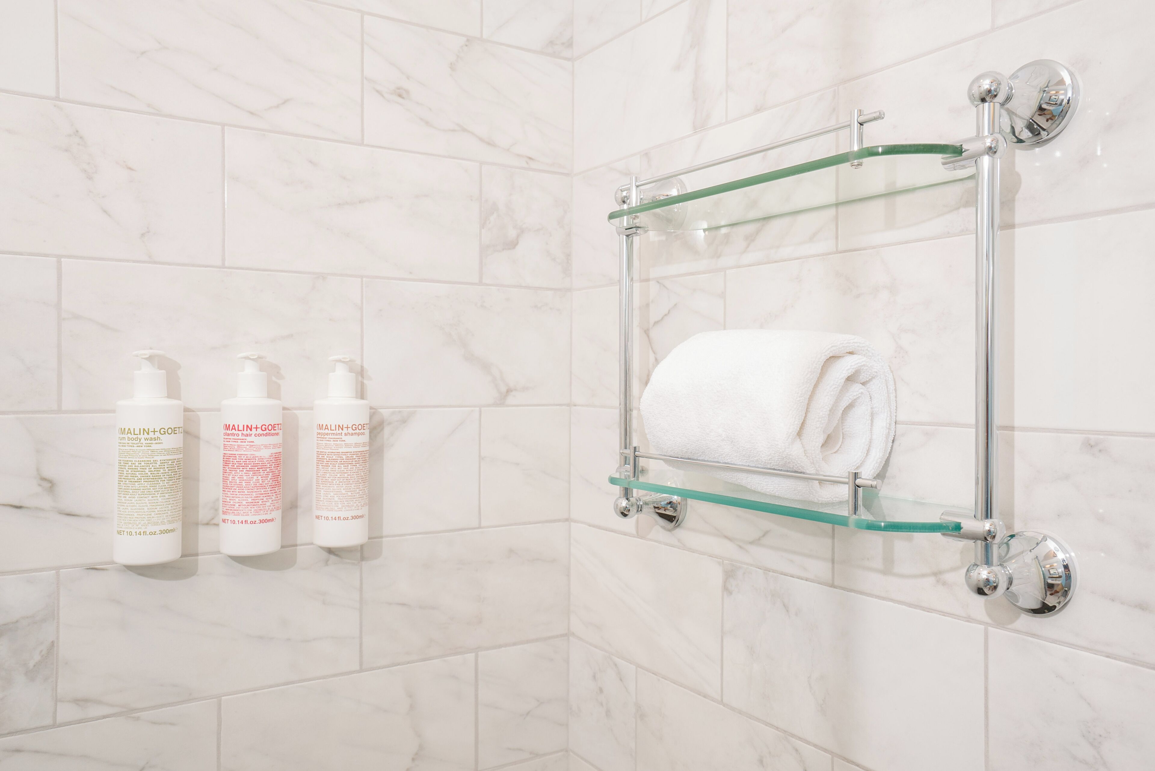 A bathroom with marble tiles, a glass towel rack with a folded towel, and three bottles of shampoo.