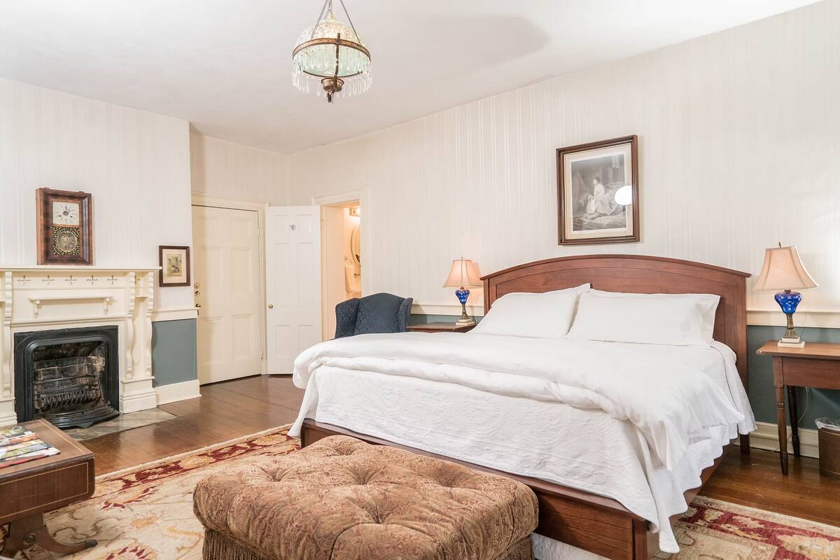 A bedroom with a large bed, a wooden headboard, a chandelier, a fireplace, and a framed picture on the wall.