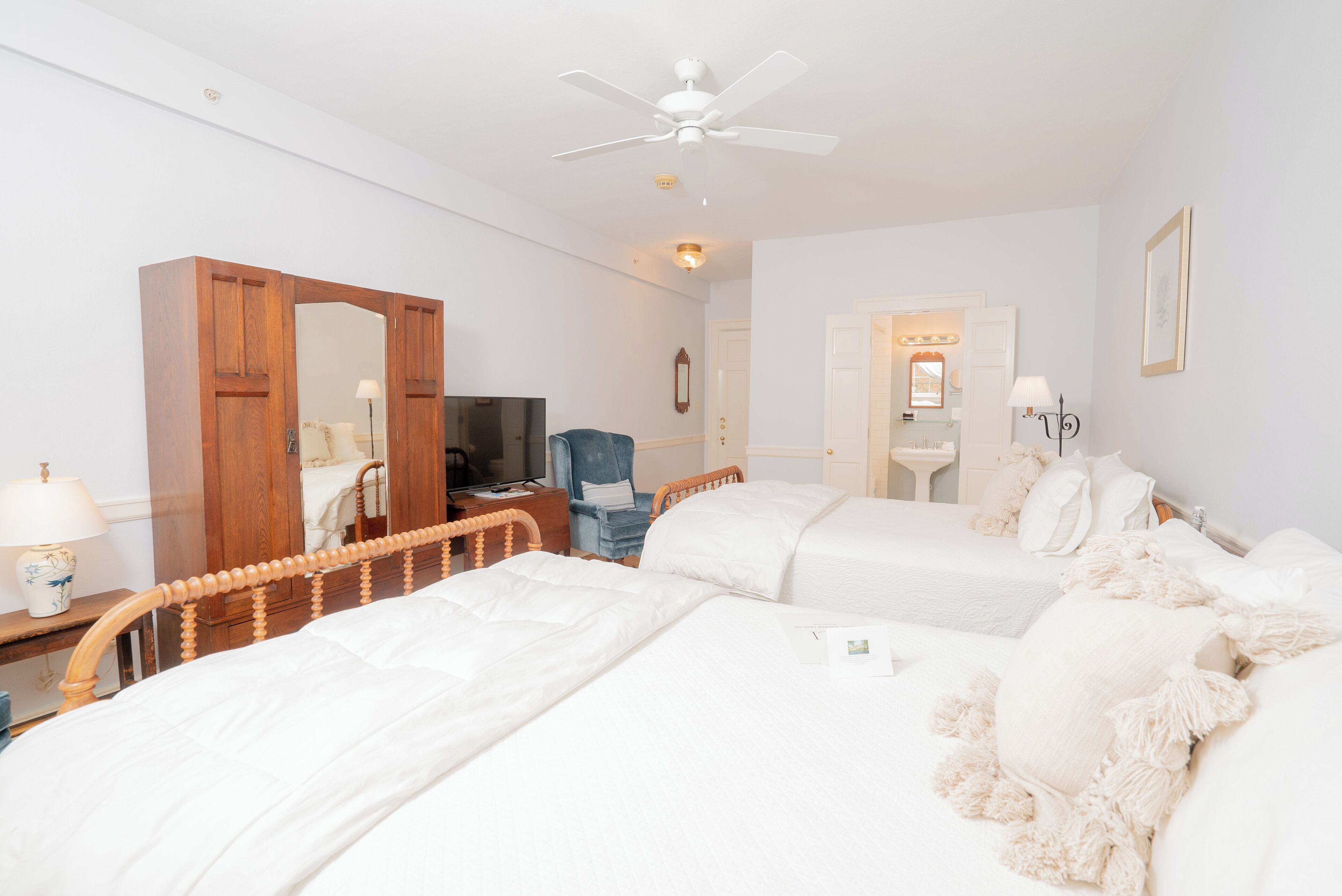 Ein Hotelzimmer mit zwei Betten, einem Holzschrank, einem Schreibtisch mit Lampe, einem Fernseher und einem Deckenventilator.