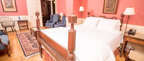 Un dormitorio con una cama de madera con dosel, un escritorio con lámpara, dos sillas y una alfombra roja.
