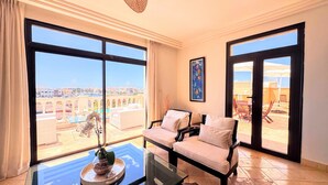Ein Zimmer mit einer Couch, einem Glastisch mit Pflanze und großen Fenstern mit Blick auf Gebäude und einen Pool.