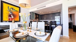 A modern dining area with a glass table set for a meal, a kitchen in the background, and a large abstract painting on the wall. - Private Rooftop Penthouse Marina Resort (Punta Cana)