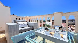 A rooftop terrace with a glass table, white cushions, and a view of the city. - Private Rooftop Penthouse Marina Resort (Punta Cana)