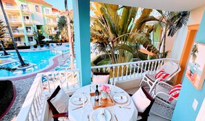 A balcony with a table set for dining, overlooking a swimming pool and a hotel building.