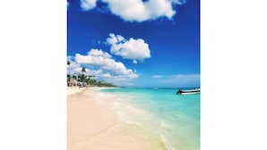 A beach with clear turquoise water, a sandy shore, and a small boat in the distance. - Walk to Beach, Shops, Dining, Pool View, Fast Wifi (Punta Cana)